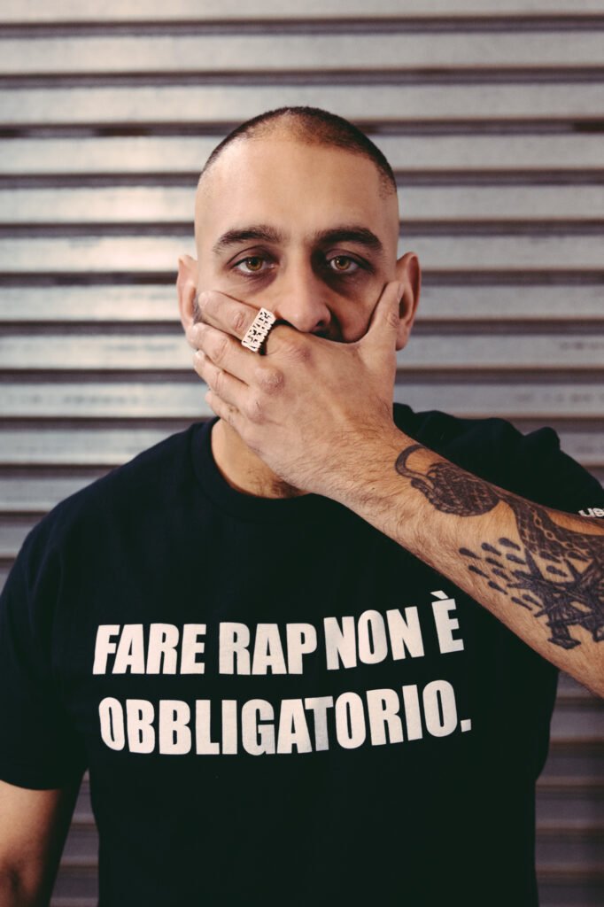 Egreen con una maglietta nera con una scritta bianca che dice "Fare rap non è obbligatorio". Ha una mano sulla bocca, sull'indice un grosso anello rettangolare di metallo, e guarda dritto in camera.
