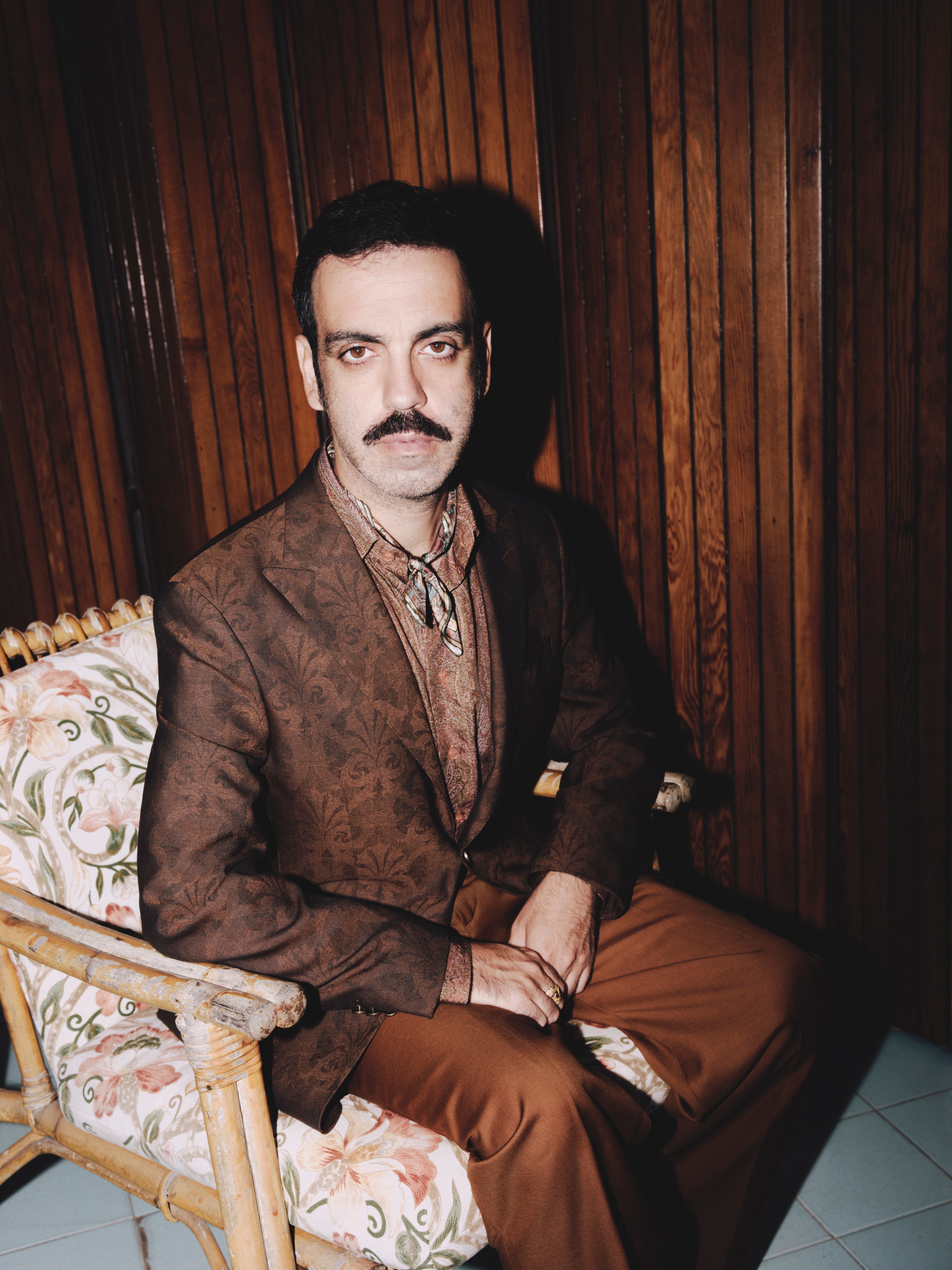 Colapesce in completo marrone, seduto di tre quarti su una sedia di bambù. Sullo sfondo delle listelle di legno in tono con il completo.