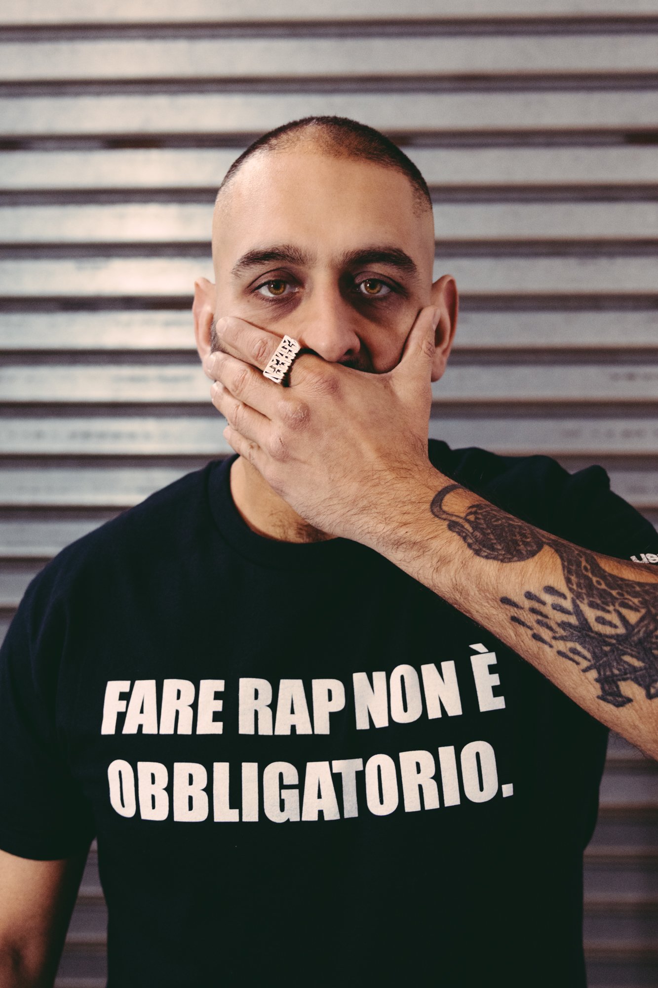 Egreen con una maglietta nera con una scritta bianca che dice "Fare rap non è obbligatorio". Ha una mano sulla bocca, sull'indice un grosso anello rettangolare di metallo, e guarda dritto in camera.