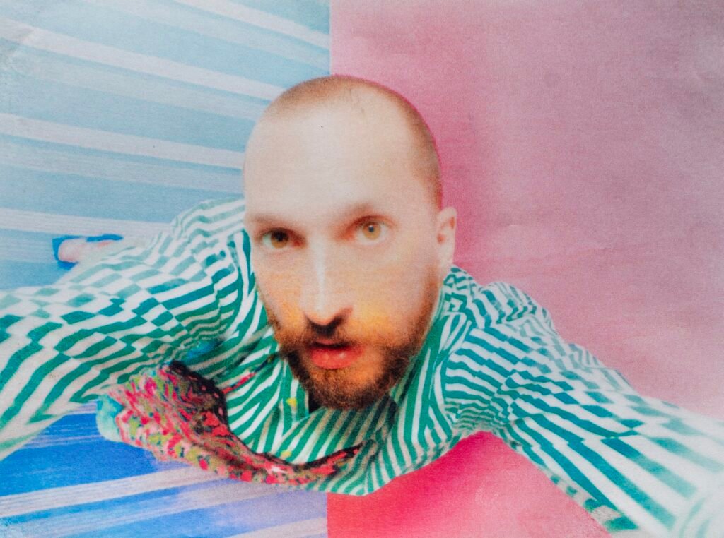 Venerus che si fa un selfie dall'alto, con una camicia a righe bianche e verdi e con sfondo un pavimento mezzo rosa e mezzo azzurro a righe bianche. la foto è un po' opaca, come venisse da una vecchia macchina foto.