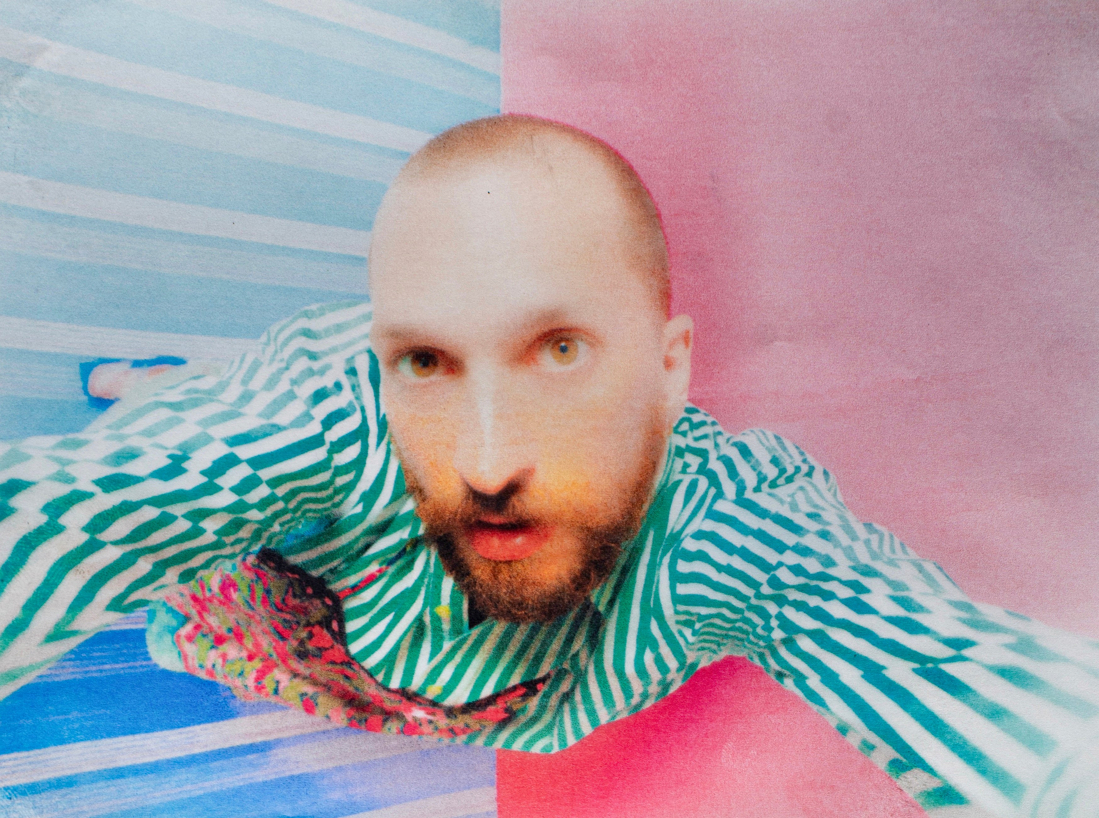 Venerus che si fa un selfie dall'alto, con una camicia a righe bianche e verdi e con sfondo un pavimento mezzo rosa e mezzo azzurro a righe bianche. la foto è un po' opaca, come venisse da una vecchia macchina foto.